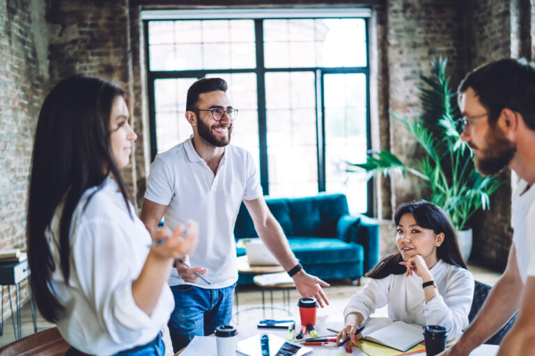 Cheerful young multiethnic coworkers gathered around table with notebooks and paper cups working together in modern loft with brick walls